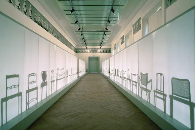 A hallway leading to a door at its end, the walls on either side of it project aligned chairs.