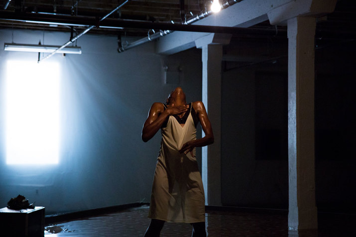 Okpokwasili leans back their head, holds one palm to their neck and pulls at their beige slip with their other hand, standing in a dimly-lit room illuminated by a lightbulb and faint light streaming in through a tarped window.