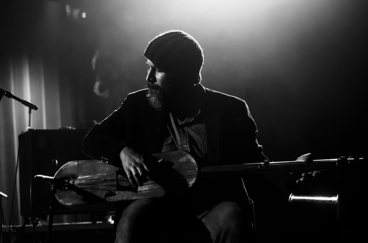 Black and white image of a figure playing a string instrument their body facing the viewer as the face sits profile. They fashion a beanie and a beard and their figure is mostly shadowed except higlights on their face from the light illuminating behind them.