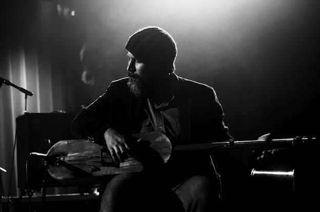 Black and white image of a figure playing a string instrument their body facing the viewer as the face sits profile. They fashion a beanie and a beard and their figure is mostly shadowed except higlights on their face from the light illuminating behind them.