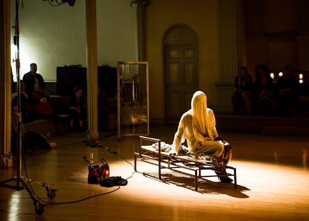 A performer sits on a bench in the middle of a room. The performer's head and upper body are covered by white draped cloth and they wear black high heels. A spotlight shines on the performer and around them, audience members sit between light blue columns, in shadow. 