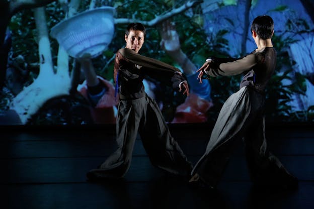 A performance still of two performers wearings billowing pants and mesh black shirts. They face each other with legs in a wide stance, each putting their weight in their right legs. Their arms are outstretched towards their left side. On the wall in the background, there is a blue hued projection on hands holding a strainer and reaching up into a tree, perhaps collecting something from it. 