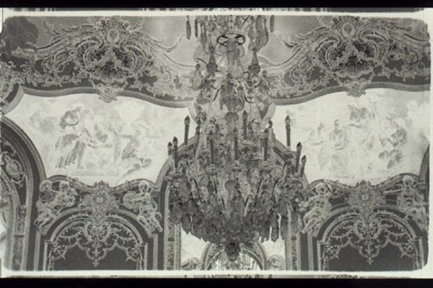 A black and white image of a decorative ceiling with panels of paintings and a chandelier. 