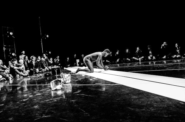 A black and white photograph of a performer positioned in the middle of the floor, nude, on their hands and knees. Around them, audience members sit on the floor and watch. The floor itself is black with a diagonal white stripe running across it. 