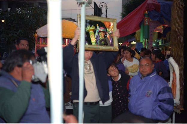 Several people crowd in a street at twilight. In the background, there is a whimsical structure made of multi colored fabric. A person in the center of the crowd holds up a gold framed image of a child in face paint next to a small green tower. 