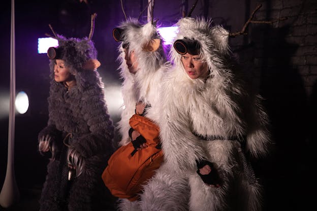 Three figures standing next to each are dressed in fur white and gray costumes with wooden antlers on the top. The figure in the middle holds an orange duffle bag. 
