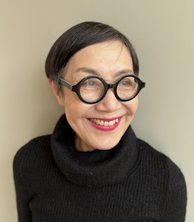 Takako Yamaguchi looks to the right of the camera and smiles showing her teeth, and in front of a beige background. She is wearing round black eyeglasses and a black knit turtleneck. 
