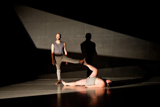 On a black walled stage cut with geometric shadows, one performer stands on one leg and extends and balances their other leg on the raised foot of another performer lying on their back. 