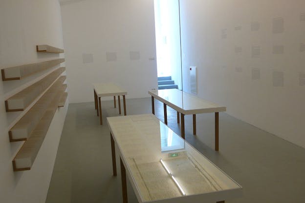 Installation view of three glass-framed tables displaying sheets of text-filled paper, a wall of stacked shelves hosting white cards, and blocks of black text on the white walls. 