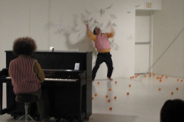 Video still of two performers wearing red and white pin-striped shirts. One performer is turned away from the camera, playing an upright piano. The other throws their hands up and looks up towards numerous blurred grey objects which surround them. On the white wall to the left of this performer, there is a thin blue drawing of four arrows pointing towards a circle in the middle. On the white floor, numerous small orange balls are scattered. 