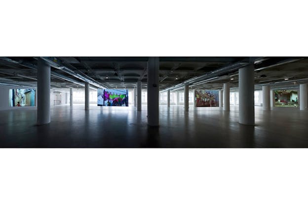 An installation image of four screens arranged within a large open space interrupted only by columns. On the far wall of the space, there are floor to ceiling windows. On the far left screen, there is an image of a child on a thin street. The next screen depicts a green and pink fabric awning. Next, there is a screen with a crowded orange train. On the far right, there is a screen depicting a white box on a dark stone background. 