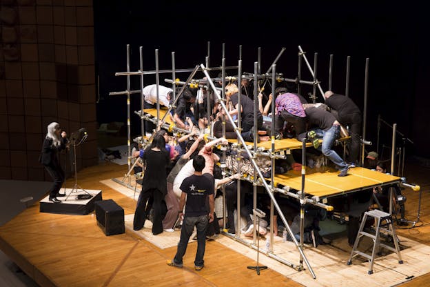 Keiji Haino stands on a raised platform on the left of the image, dressed in black and holding a microphone. A swarm of performers climbs under, through, and on top of a structure made from metal poles and yellow platforms, crowding each other.