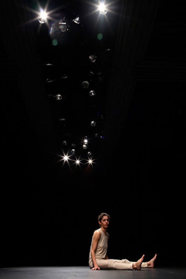 A performance still of Maria Hassabi sitting with her legs out in front of her on the black floor of a stage. She loosk out towards the audience with a pained expression on her face. She wears all tan and had her hair pulled into a bun. Above her, nurmerous stage lights shone slightly, illuminating her in the otherwise dark space. 