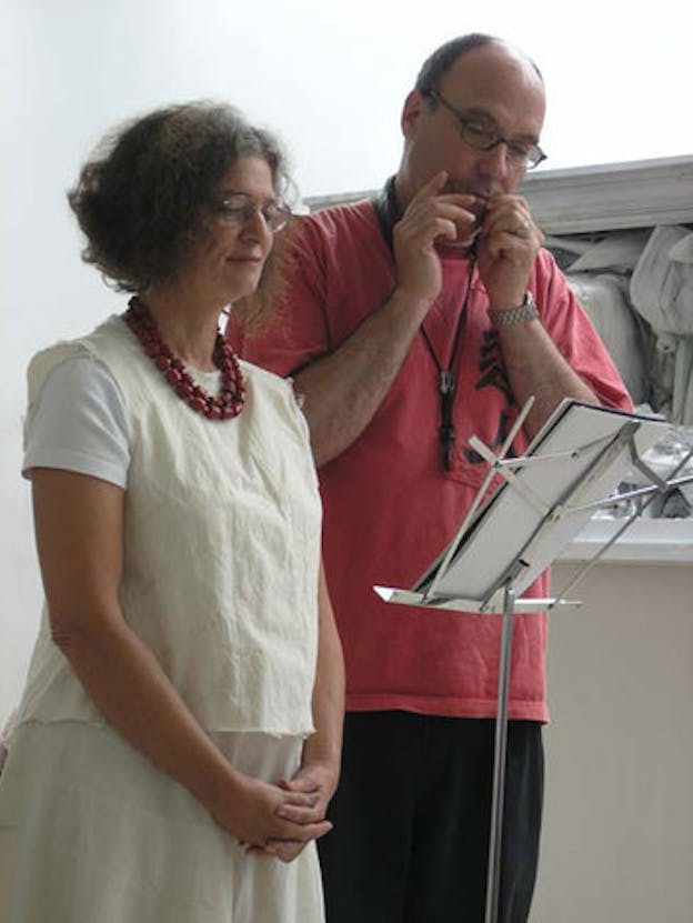 Two people looks down at a music stand together while standing in front of a white wall with a large relief sculpture. 