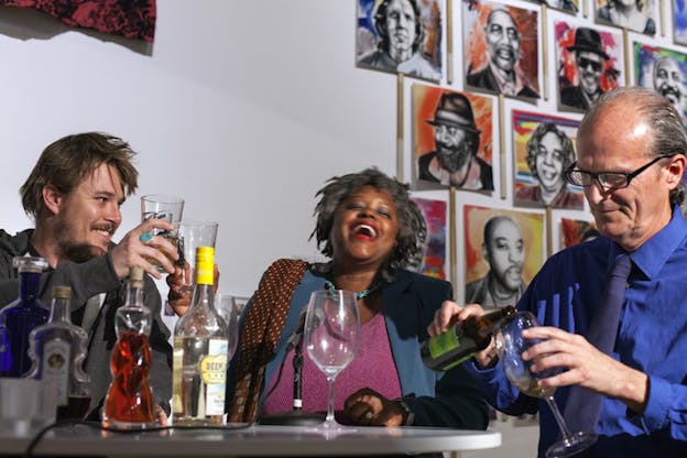 Three people sit on a table laughing with glasses and bottles in front of them. Behind them on the wall, a collage of various paintings.