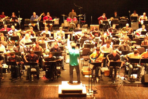 Aerial view of a conductor gesturing to a large assembly of seated musicians playing guitars and in the center, one person playing red drums. 