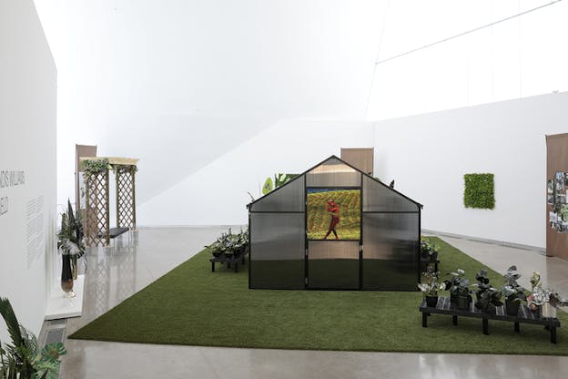 A makeshift green field stands in the middle of a white space. Above it a house with blurred glass panels has the image of a red figure in a flower field. Surrounding it wooden panels and glass vases with leaves.