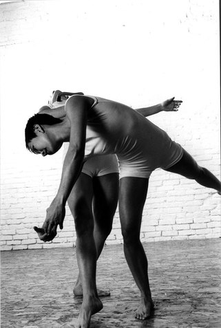 Two figures dressed in white perform together. The figure on the front closest to the viewer raises one leg and leans forward while the figure in the back leans their upper body backwards. 