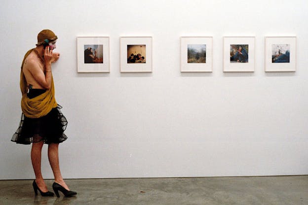 A person wearing black pumps, a sheer black skirt, and a tan drape top speaks on the phone while leaning against a white wall displaying five small framed images. 