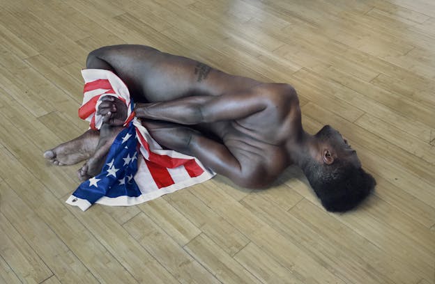 A performance still of the back of Carlos Martiel as he lays on a wooden floor. His arms and legs are bound together, behind him, with an american flag. A script tattoo is visible on his hip. 
