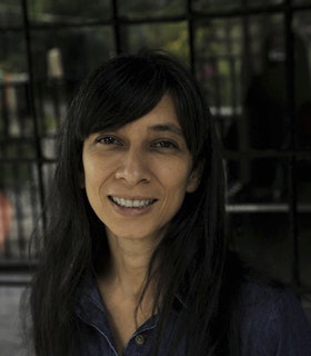A portrait of Regina José Galindo smiling, with long dark straight hair and side swept bangs, wearing a blue denim collared shirt.