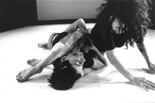 A close up black and white image of two performers in a bare white space. One performer sits on the other performer who is laying down. The two performers look intently at each other. 