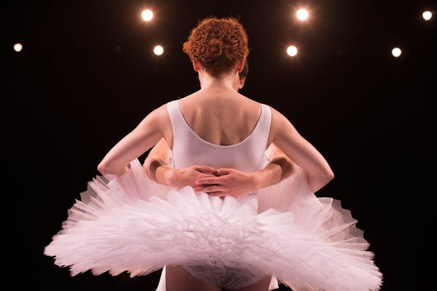 Low angle shot of a ballet dancer beneath bright stage lights, their white tutu fanning out, another performer's hands supporting their back.