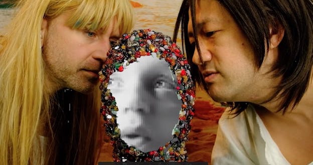 Sean McElroy and Tei Blow lean in from either side to smell a collaged black and white image of a baby, face covered in a hood of pills and supplements. McElroy is wearing a long, straight blond wig with bangs, and Blow is wearing a shoulder length dark brown wig with long bangs. Both gaze at the head between them.