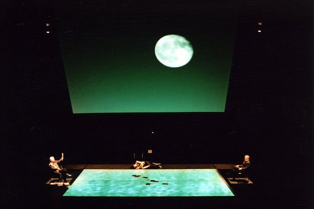 A green projection with the moon on a far wall and on the ground. Two people sit in front of the shorter sizes of the projection, while a third lays down along the longer side.