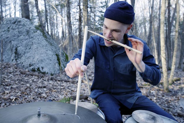 A person dressed in dark blue with a beanie, seated behind drums in a forest hold a drum stick between their teeth.