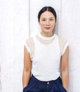 Anna Sew Hoy stands against a white wall looking directly at the camera. She wears a white, short sleeve top with blue trousers. Her hands are tucked into her pockets.