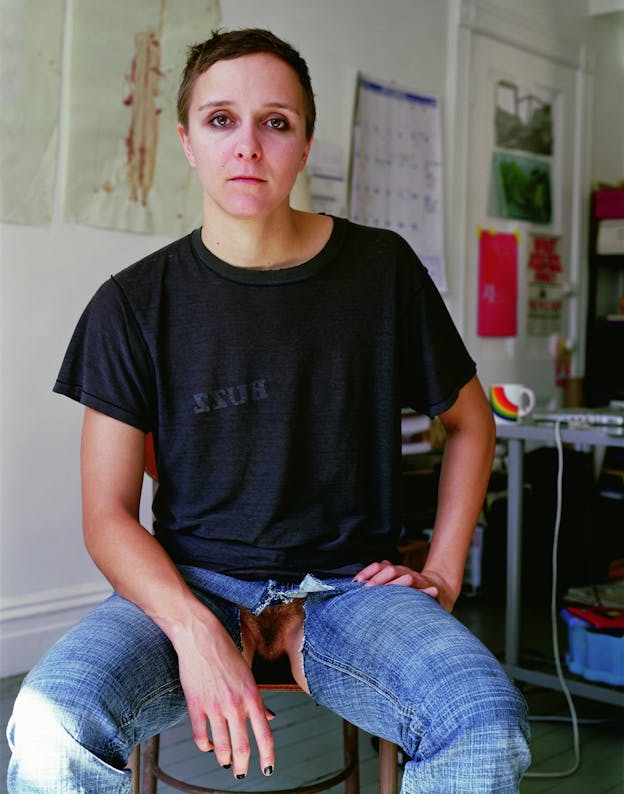 A photograph of someone sitting on a chair, looking directly at the camera. They are wearing a black t-shirt that has 