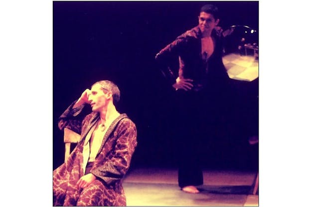 Two performers are in a dark space. The performer on the left wears a marbled dark robe and sits in a chair, with their head in their hand and looking ahead of them at something beyond the frame to the left side. The second performer stands in shadows. They wear a red and purple shirt, a pendant, satin pants and have bare feet. With their left arm, they hold a ciurcular mirror towards the other performer and smile. 