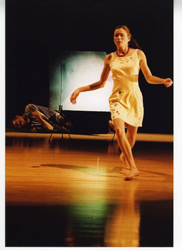 A performer wearing a white dress appears to be in the middle of running. Both of their arms are held open and bent at the elbow and their legs are bent at the knee, one leg in front of the other. Behind them, a performer lays curled up on the ground. The background is all black except for a blurred turquoise projection.