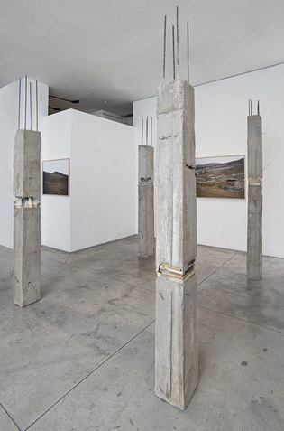 Four building concrete pillars are placed on a concrete floor to form a rectangular shape. The installation is situated in an interior spaces of white walls with framed works depicting landscape attached to them.