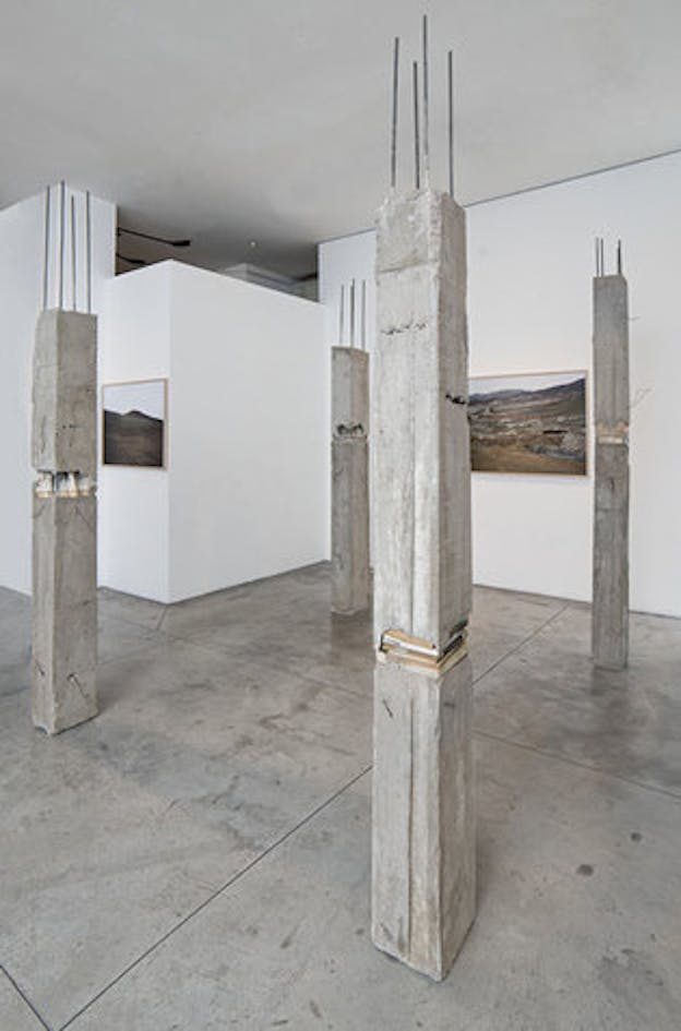 Four building concrete pillars are placed on a concrete floor to form a rectangular shape. The installation is situated in an interior spaces of white walls with framed works depicting landscape attached to them.