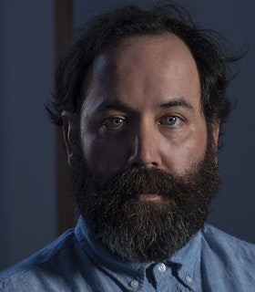 Nathan Young looks directly at the camera in front of a dark blue, out of focus background. He is wearing a light blue linen button down shirt with white buttons. 