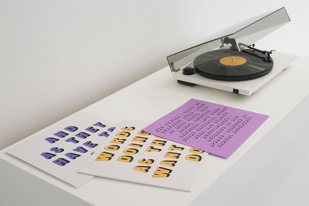 A white shelf in a gallery with two layered record sleeves and an active record player on it. One sleeve is printed in purple with the (partially obscured) words: 