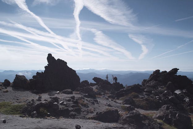 A video still of a rocky terrain. A far away person is visible as they lean against a small tree in the middle of this terrain. The sky is bright blue sky and criss-crossed with thin lines of clouds. In the background, a blue hued mountain range is visible. 