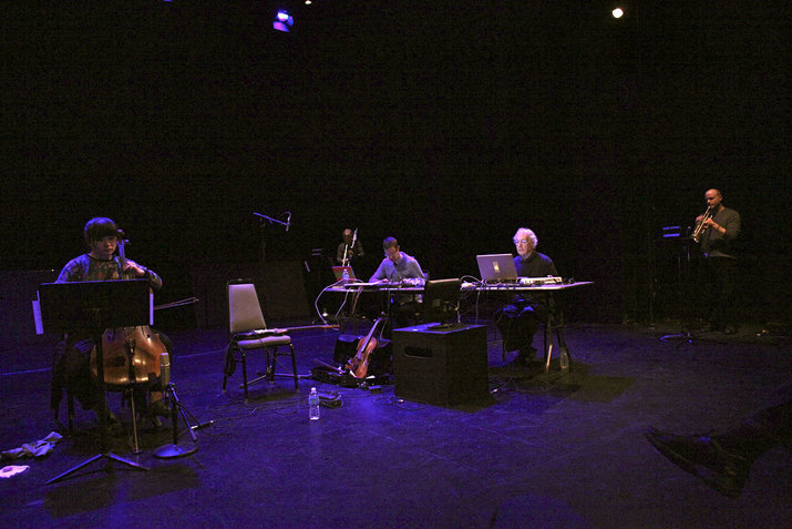 Performers seated on chairs on a purple lit stage. The performer to the left plays a string instrument as two in the middle face them behind computer screens situated on desks. The performer furthest from the viewer remains standing and plays a woodwind instrument.    