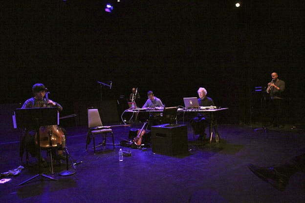 Performers seated on chairs on a purple lit stage. The performer to the left plays a string instrument as two in the middle face them behind computer screens situated on desks. The performer furthest from the viewer remains standing and plays a woodwind instrument.    