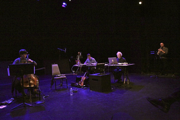 Performers seated on chairs on a purple lit stage. The performer to the left plays a string instrument as two in the middle face them behind computer screens situated on desks. The performer furthest from the viewer remains standing and plays a woodwind instrument.    