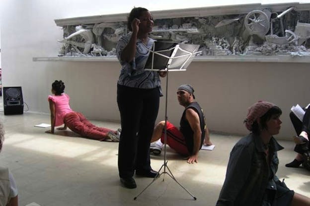 One person standing in the center looks down at the music stand while a few others are on a white floor in various positions. Behind them, a large relief sculpture is hung on a white wall.