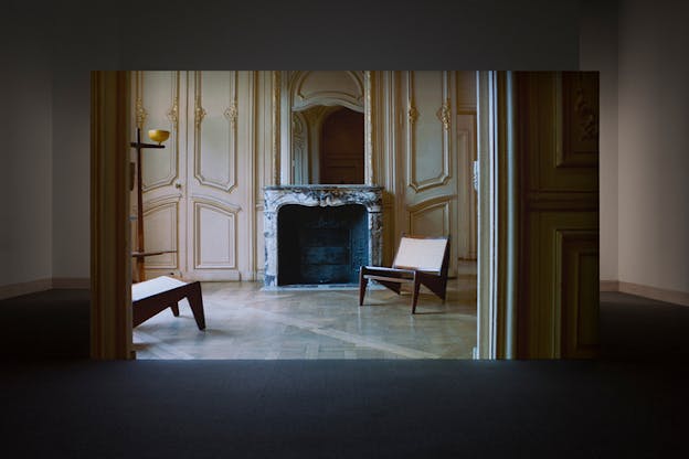 A projection of a room with white walls outlined with gold. In the middle sits a fireplace with a black and white marbled design, next to it is a low chair.