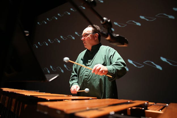 Close-up of Winant holding percussion mallets, performing in front of black screen projecting geometric blue shapes.