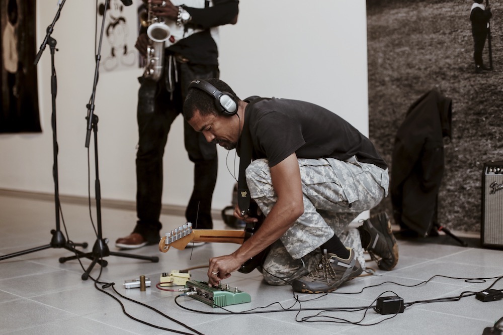 Morgan Craft crouches to adjust the pedal setup in a gallery space. He is facing left, holding his guitar at his side with his right arm while his left hand is adjusting dials. Amp and microphone wires trail around him. Another performer holding a saxophone is visible from the waist down in the middle ground. Morgan craft is wearing headphones over his ears, a black t-shirt, and green camouflage cargo pants with brown hiking shoes. 