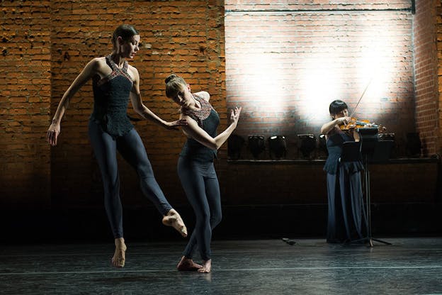 On a dimly lit stage with a brick wall backdrop, one person dressed in gray leaps pointing one foot forward and curving their right arm towards another identically dressed performer who is tilting their head and shoulders towards the other dancer and bending their knees slightly. To their right a performer plays the violin. 