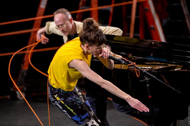 Joanna Kotze performs on a brightly lit stage with of Ryan Seaton who is seated at a black grand piano behind her. There are red ladders visible in the background out of focus. Kotze hunches forward and speaks into a microphone on a stand in her left hand, her right arm is stretched forward and pointing down. Seaton's head, shoulders, and arms are visible behind her as he faces the camera leans to the left, using his right hand to yank an orange electrical cord. Kotze is wearing a yellow sleeveless top and black and grey abstract patterned leggings.