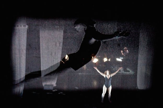 Performer in a black leotard raises their arms above their tilted back head on a dimly-lit stage in front of a projection depicting a transparent shadowy dancer suspended in front of a conductor conducting. 