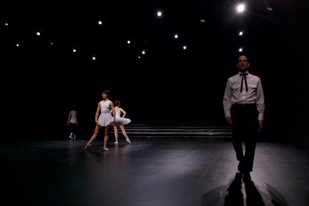 Performer wearing a black suit and tie stands in the front right of the stage and in the back left, dancers dressed in white point their feet in side tendu.